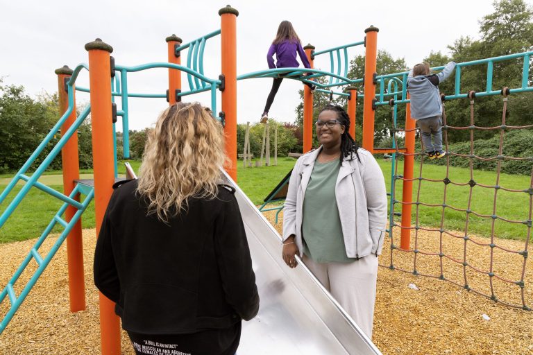 Twee vrouwen praten, kinderen spelen in speeltuin.